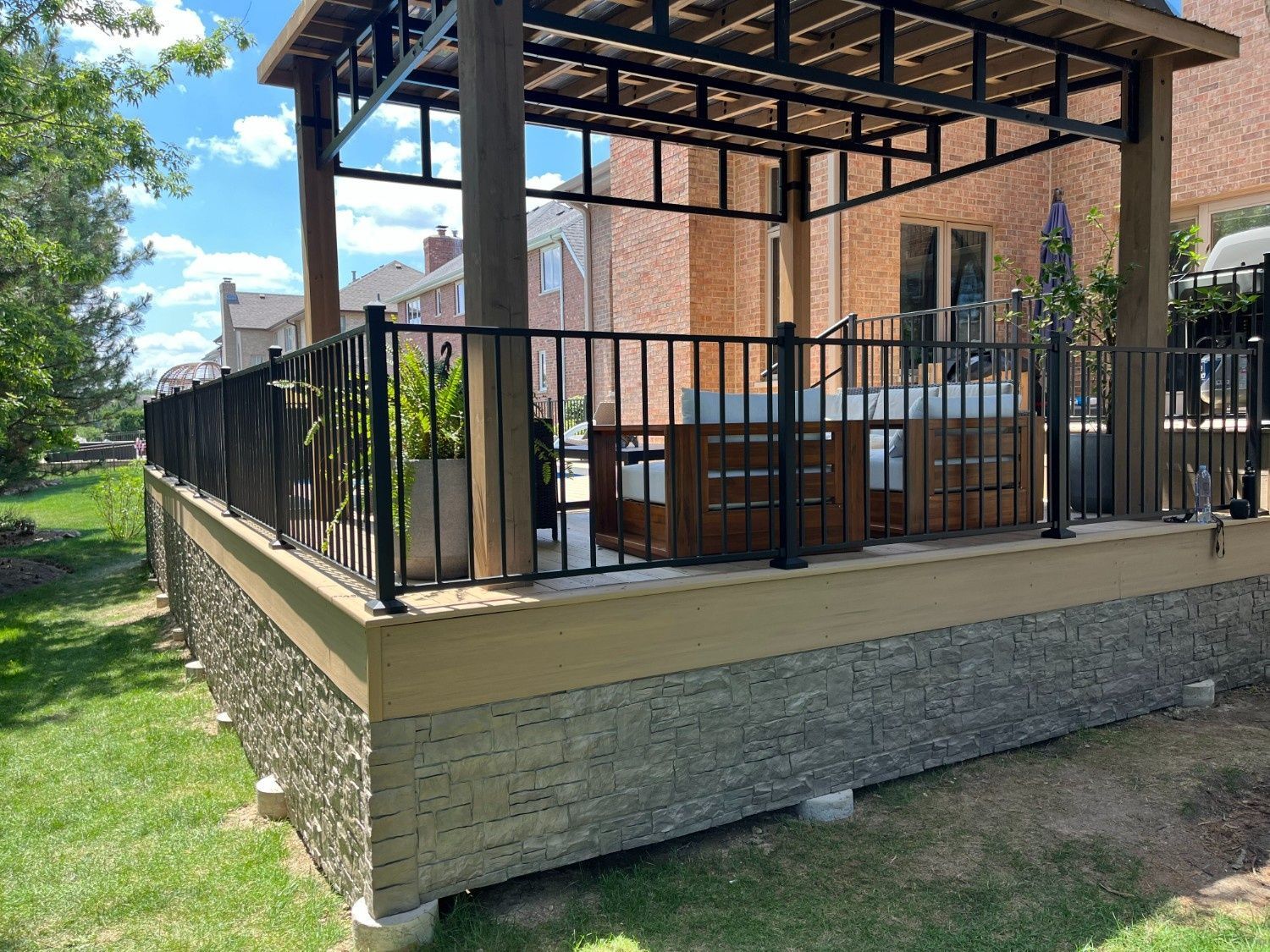 Patio deck with black railing, pergola, and outdoor seating beside a brick house.