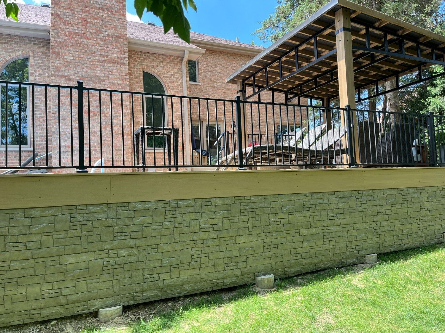 Brick patio with a metal fence, lounge chairs, and a covered pergola beside a grassy slope.