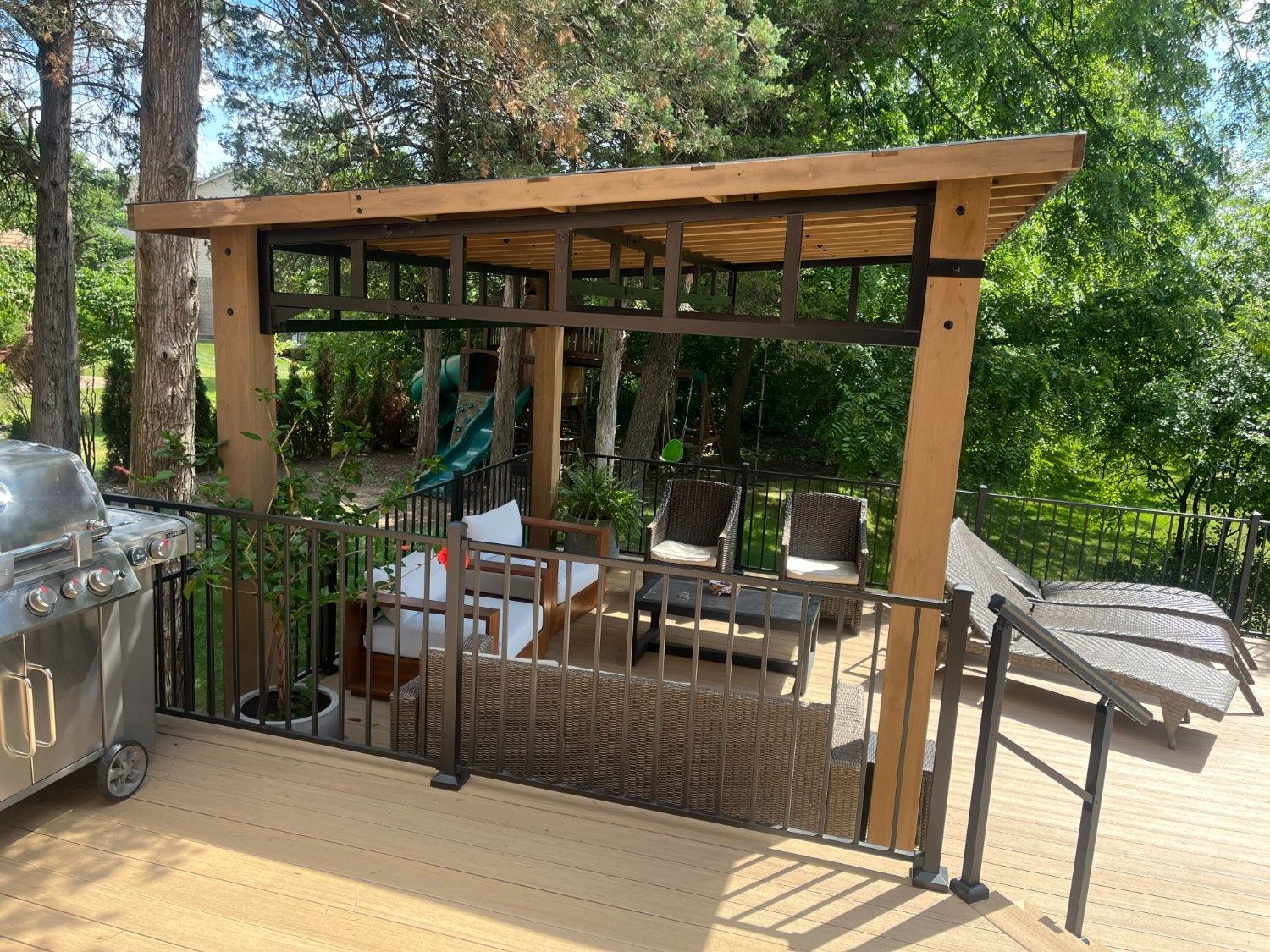 Wooden patio gazebo beside a grill, with outdoor chairs and greenery in the background