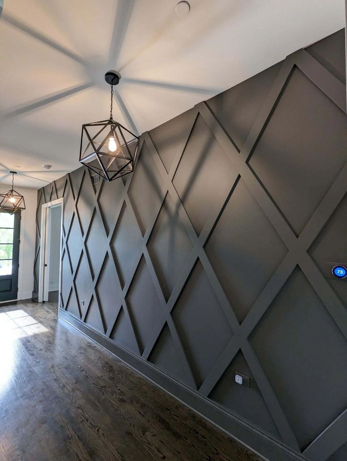 Hallway with dark diamond-pattern accent wall, pendant lights, and wood floor lighting from window