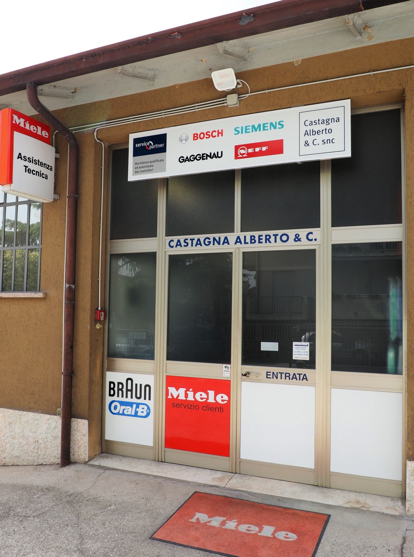 Negozio con vetrina e insegne di marca per la riparazione di elettrodomestici, tra cui i loghi Miele e Braun.