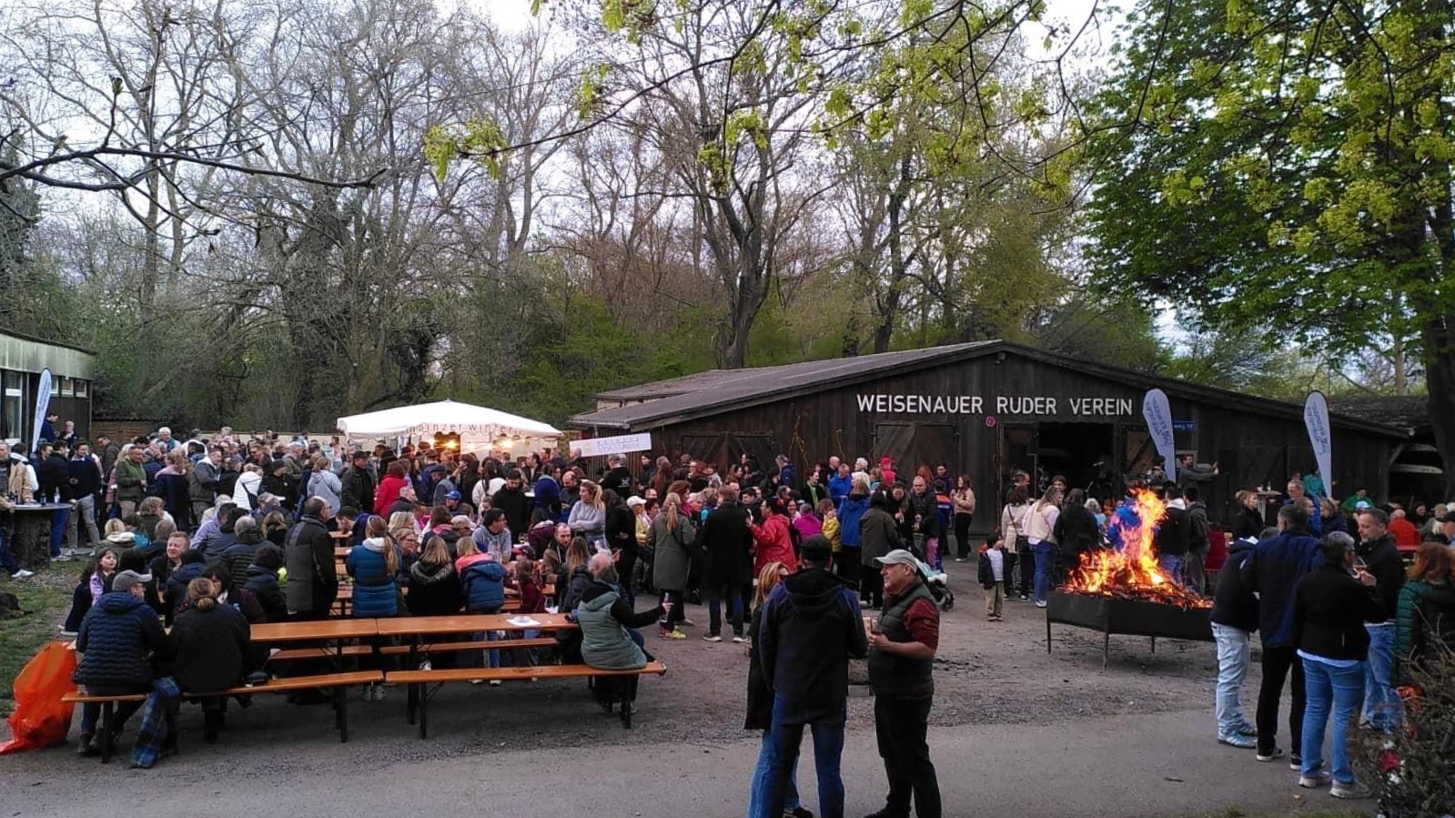 Am Abend versammelt sich eine Menschenmenge im Freien in der Nähe eines Holzgebäudes und einer großen Feuerstelle;