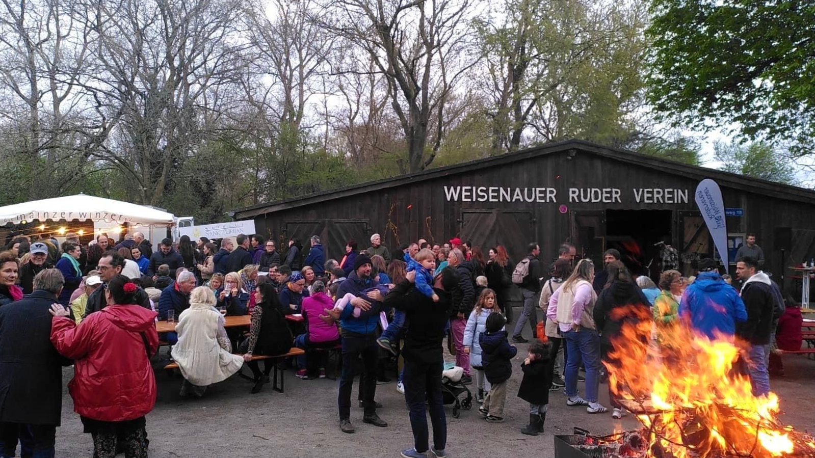 Eine Menschenmenge versammelt sich im Gebäude zu einer abendlichen Freiluftveranstaltung mit einem großen Lagerfeuer.