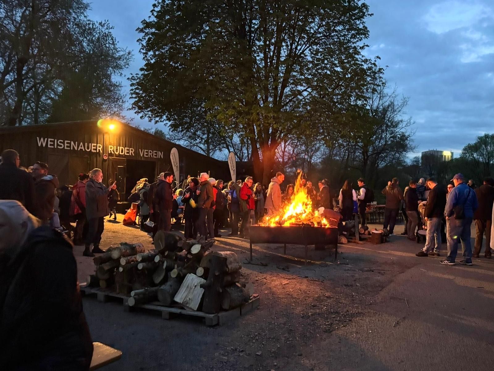 In der Abenddämmerung versammelt sich eine Menschenmenge um ein Lagerfeuer in der Nähe eines Gebäudes.