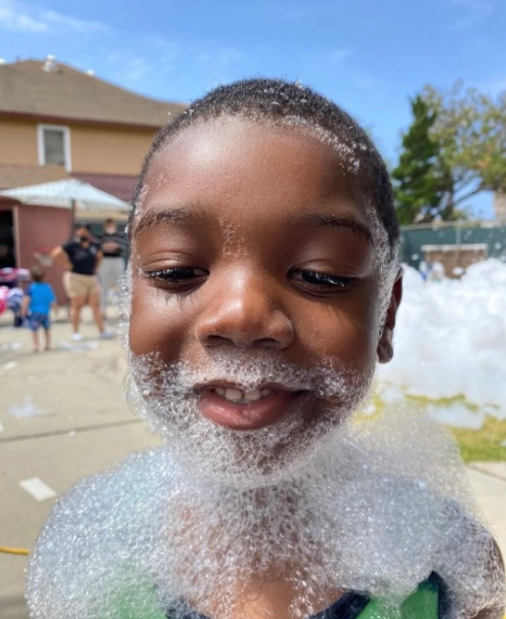 A young boy with soap bubbles on his face is smiling.