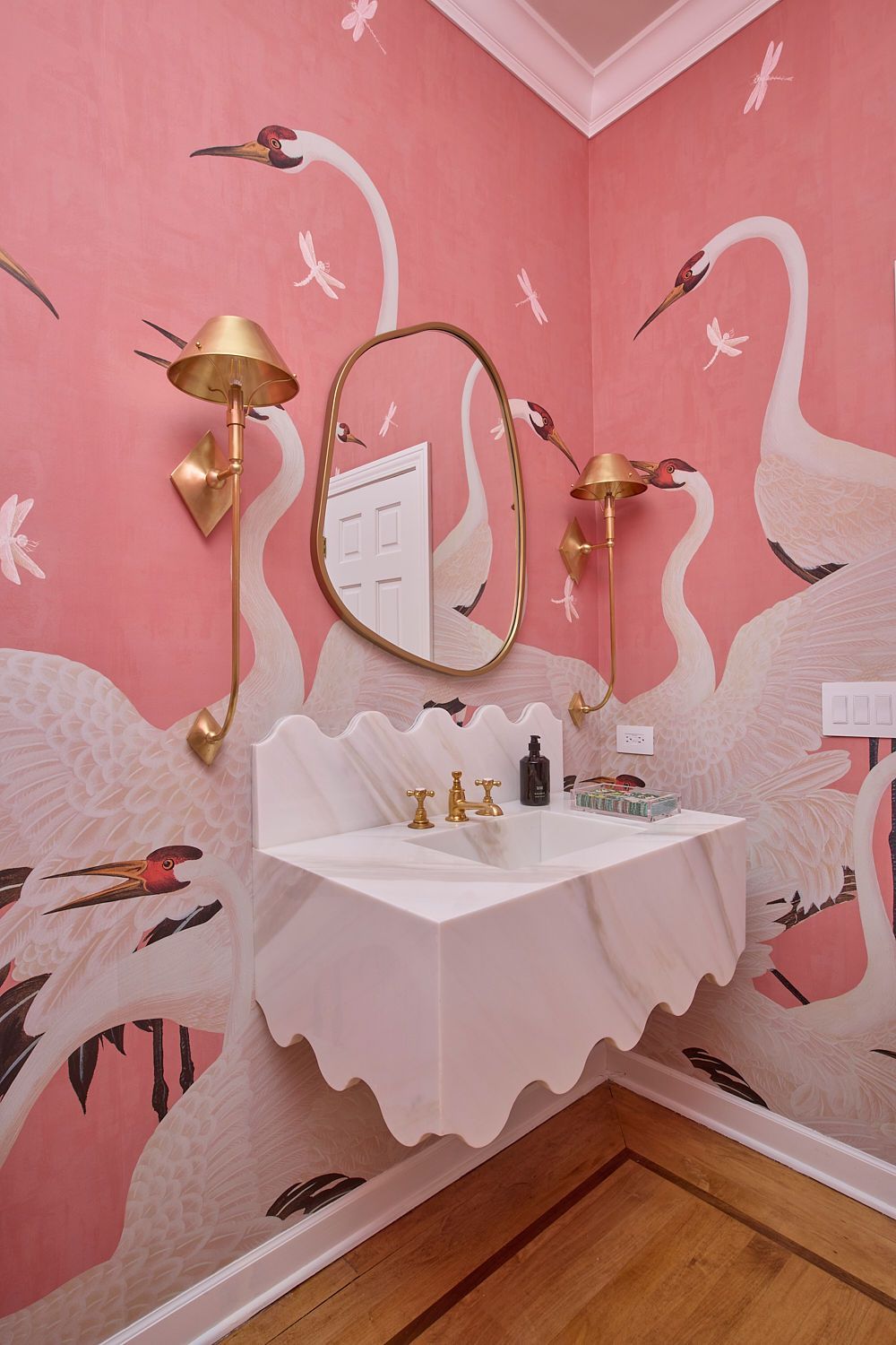 Pink wallpapered bathroom with crane mural, wavy-edged sink, gold fixtures, and ornate mirror.