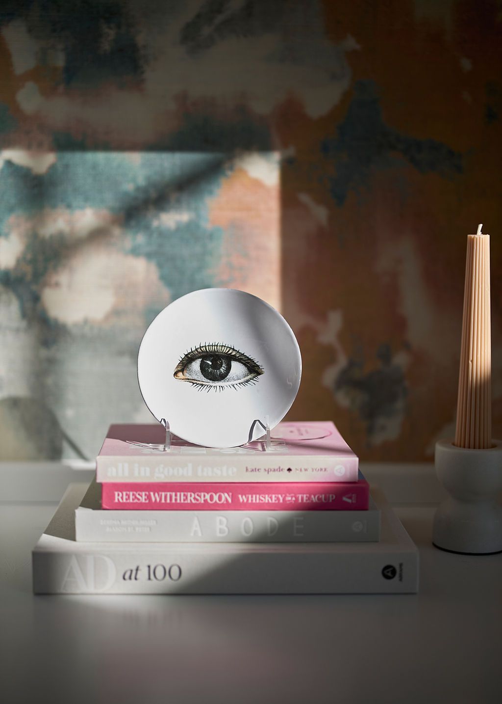 Eye illustration on a white disc atop stacked books. Pink and white books with a lit candle, against a patterned wall.