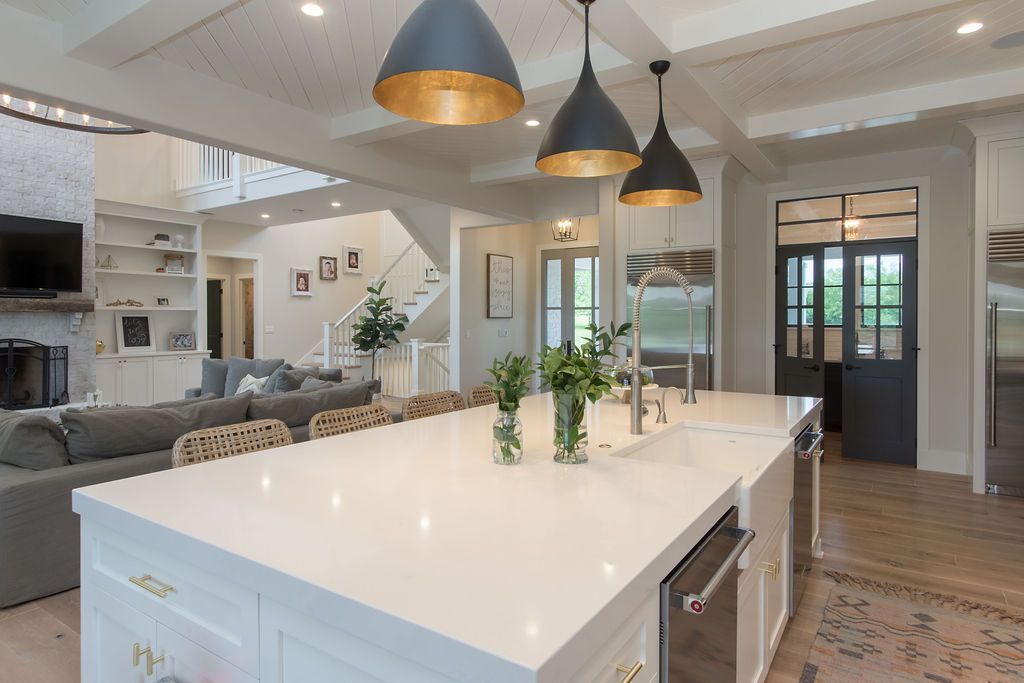 Spacious white kitchen with island, pendant lights, and stainless steel appliances. Adjacent living area.