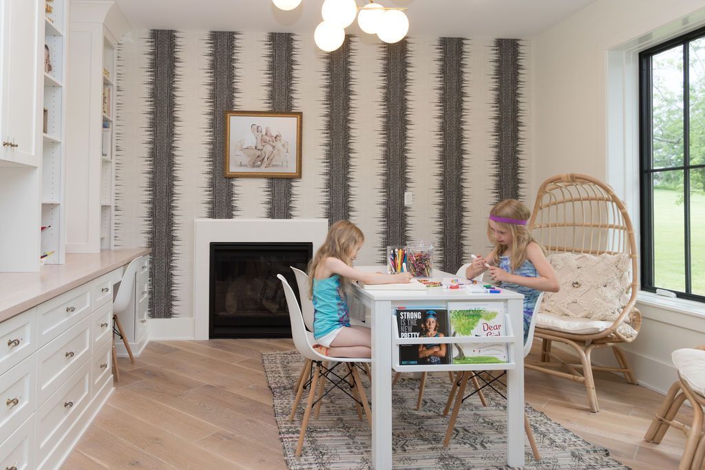 Two children coloring at a white table in a room with a fireplace and patterned wallpaper.