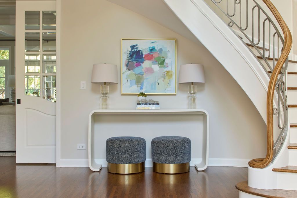 Entryway with white console table, abstract art, two gray stools with gold bases, and a curved staircase.