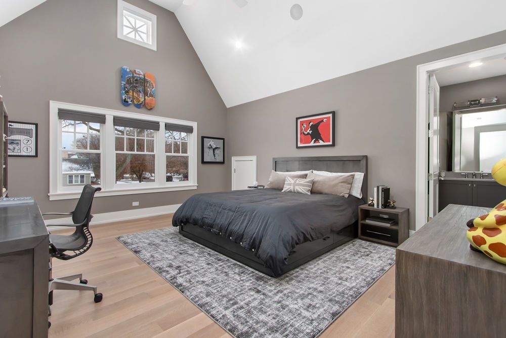 Gray bedroom with high ceiling, bed, desk, artwork, and bathroom entrance.