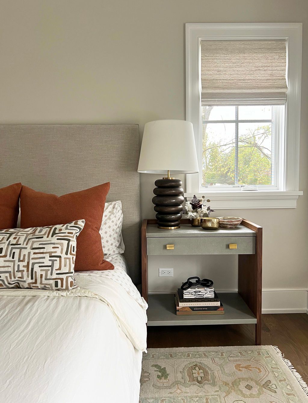 Bedroom with neutral tones, nightstand with lamp, bed with orange and patterned pillows, window with woven shade.