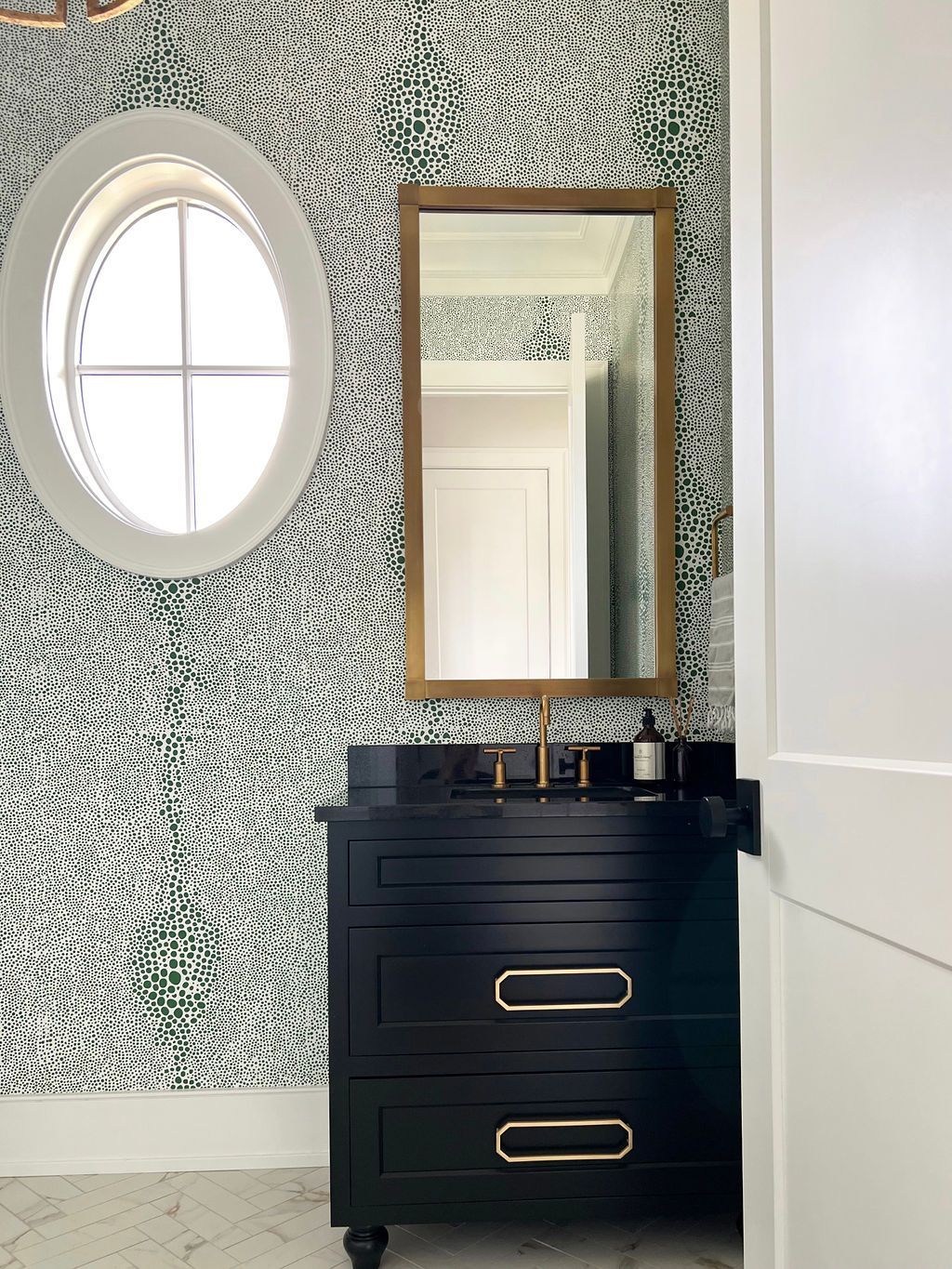Black vanity with gold accents, mirror, and oval window in a bathroom with patterned green and white wallpaper.