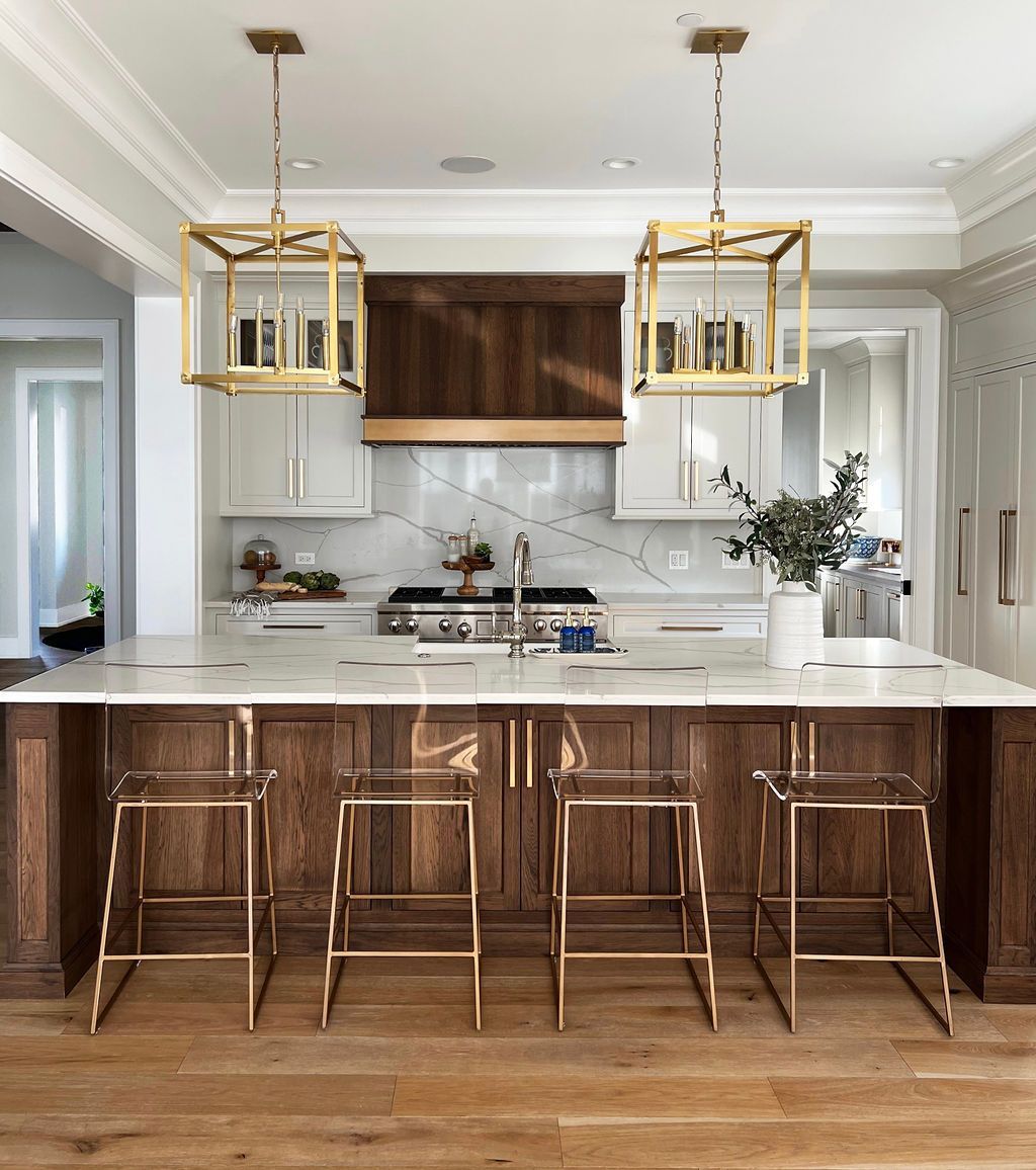 Modern kitchen with a dark wood island and gold-framed pendant lights over a marble countertop, with clear acrylic stools.
