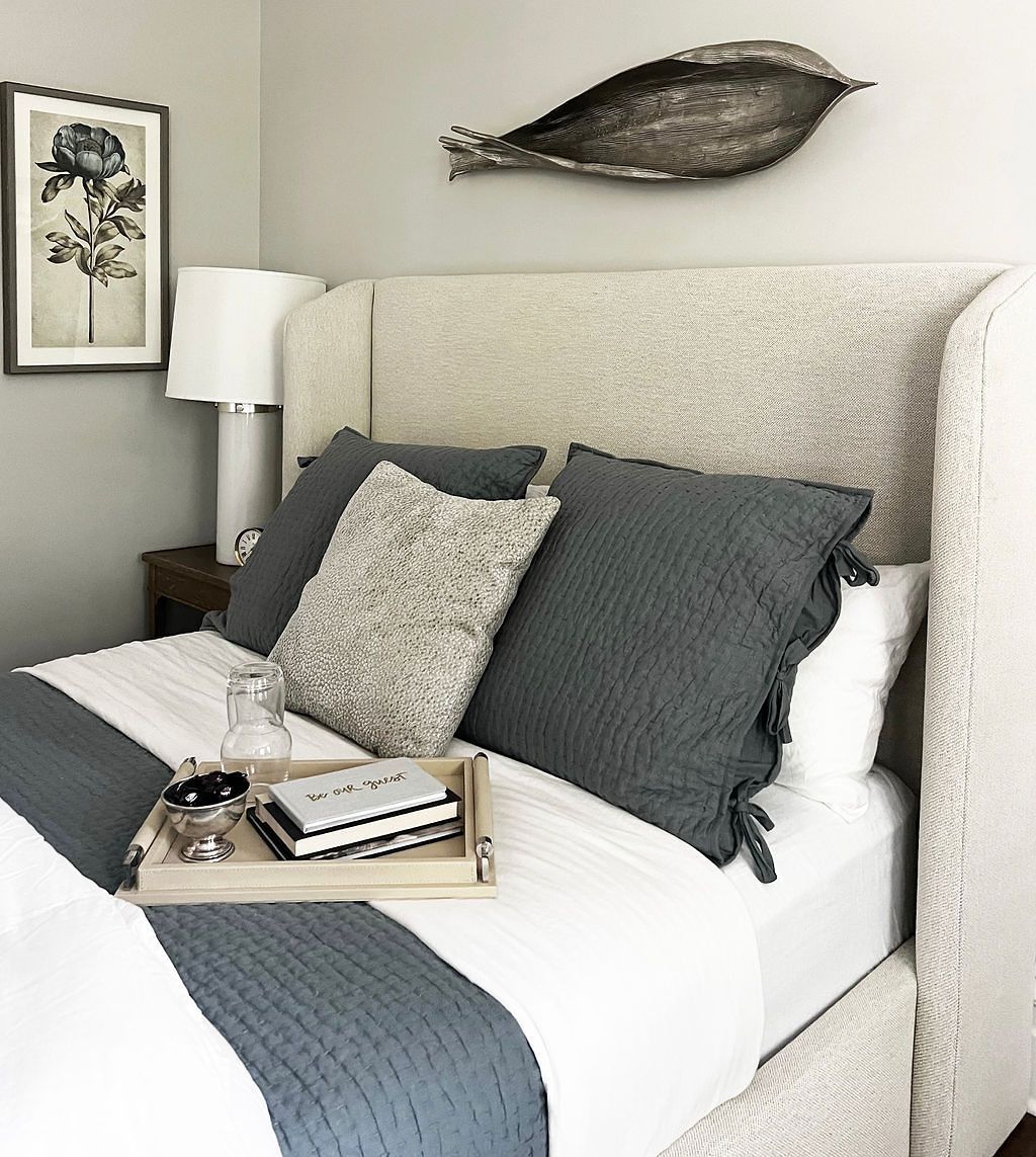 Bedroom with a bed, headboard, pillows, bedding, artwork, and decor in neutral colors.