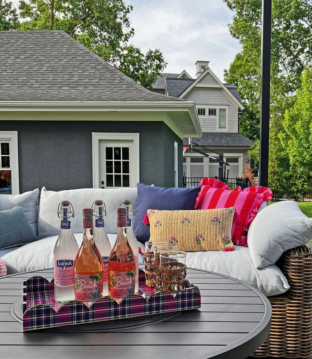 Outdoor patio with bottles of rosé on a plaid tray, pillows on a wicker couch, with houses in the background.