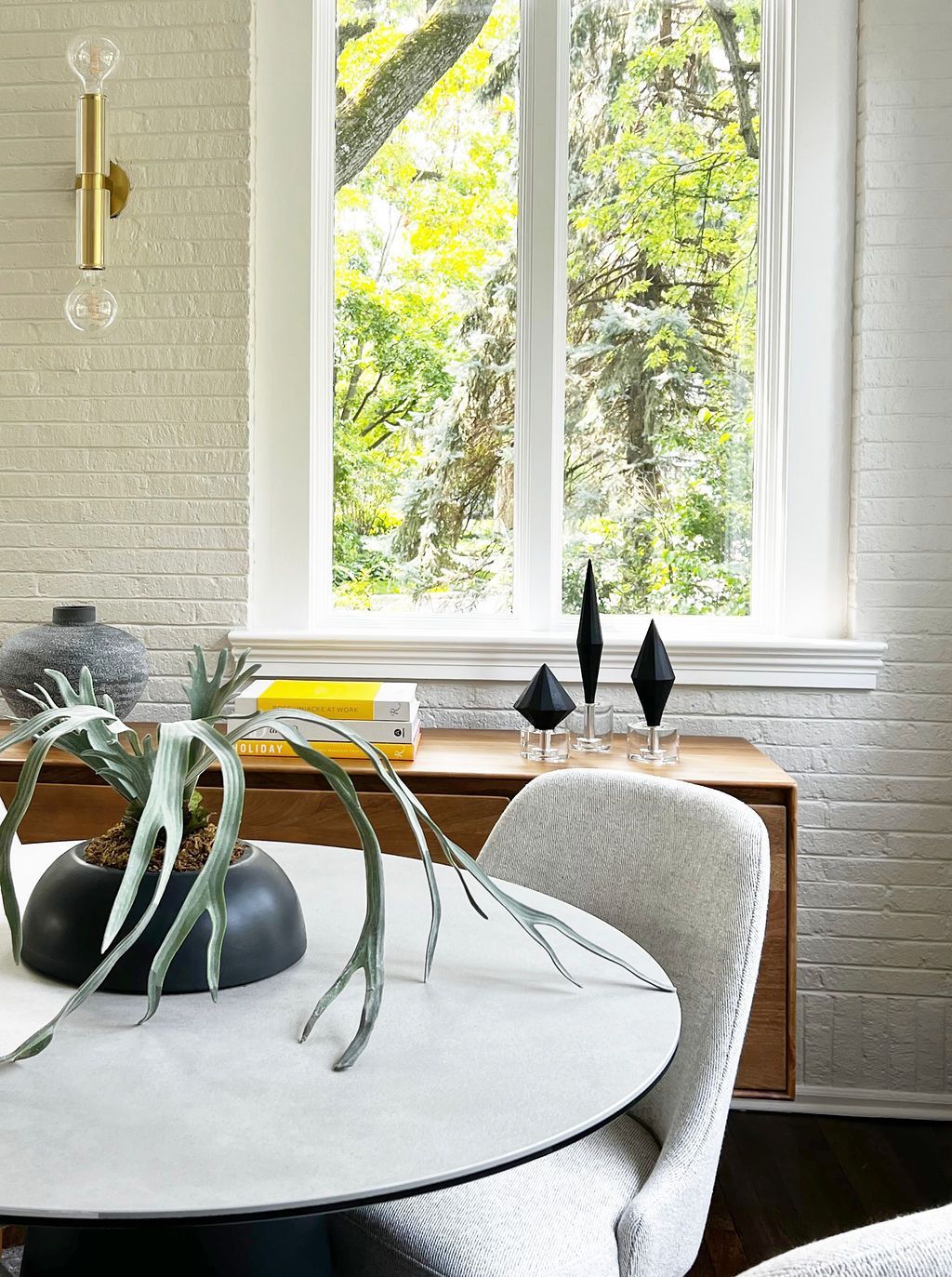 Dining room with round table, chair, and view through a window. Green foliage visible outside.