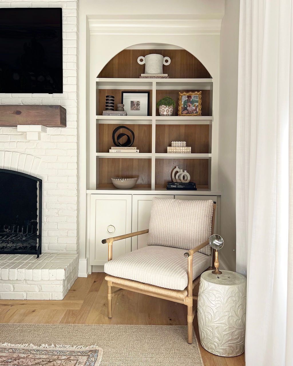 Built-in bookcase with arched top, containing decor. A patterned chair sits near a side table. White brick fireplace nearby.