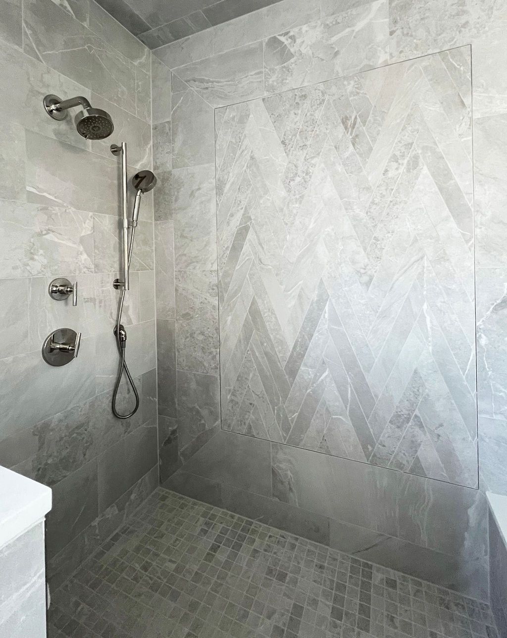 Gray tiled shower with multiple showerheads and a herringbone pattern accent wall.