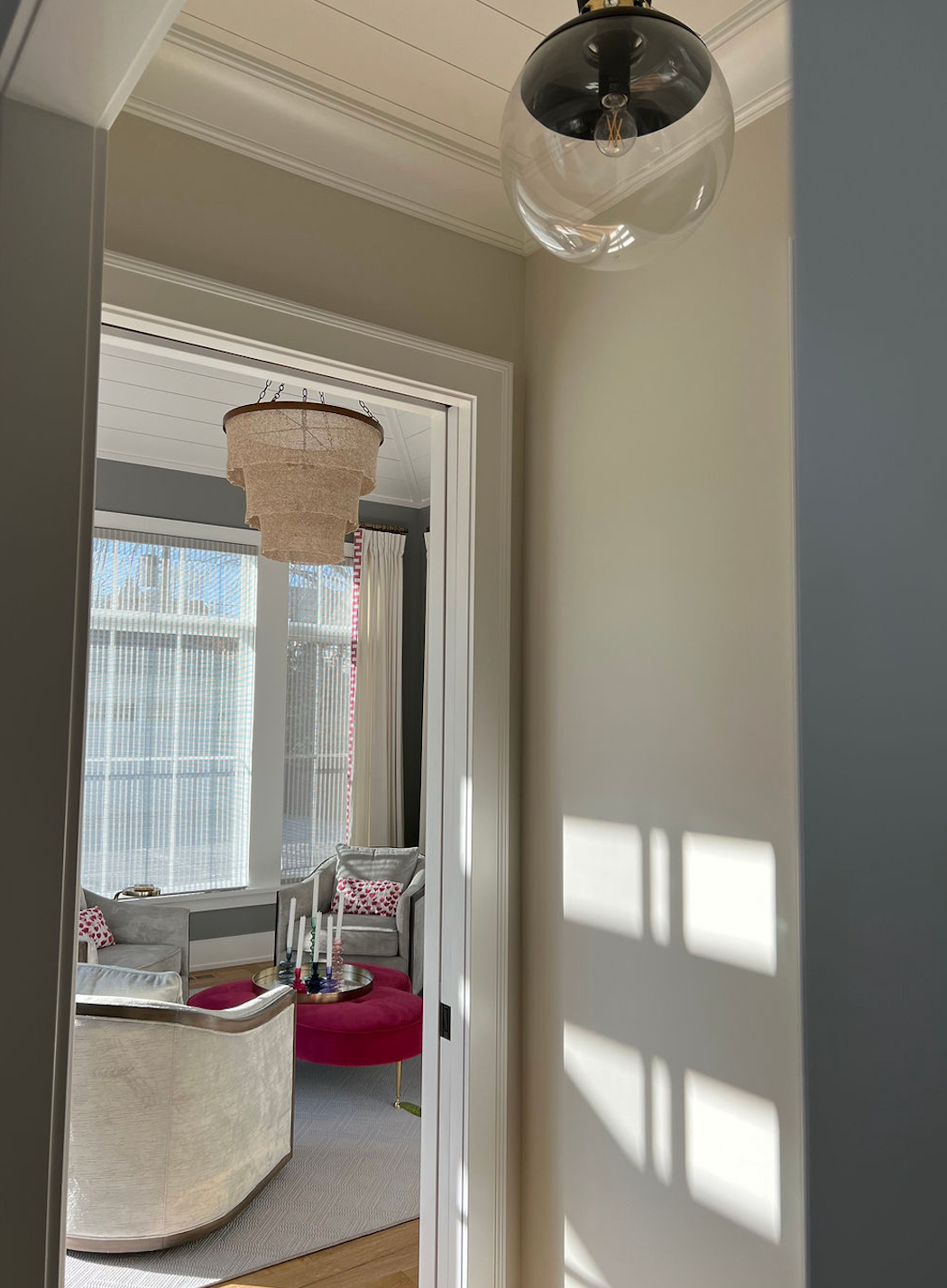 Hallway with light fixture and view into a room with chandelier, sofa, and pink ottoman.