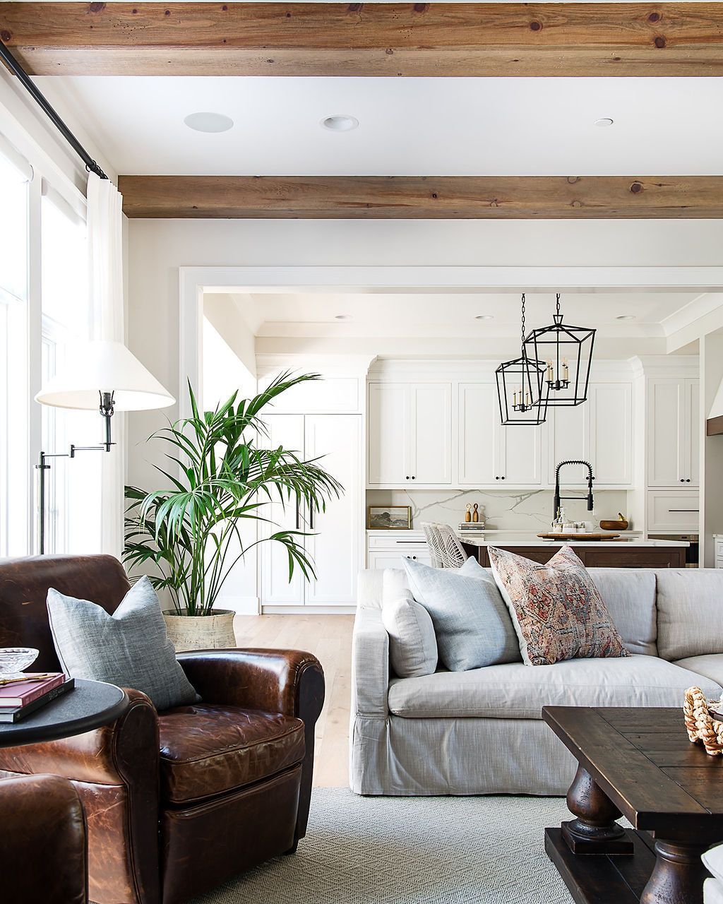Living room with leather chair, light-colored sofa, palm plant, wooden beams, and open kitchen.