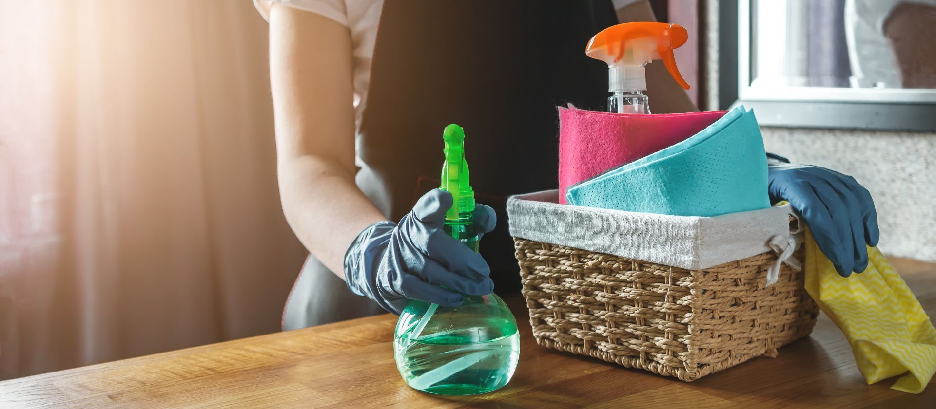 A person wearing blue gloves holds a spray bottle next to a wicker basket containing cleaning cloths and supplies.