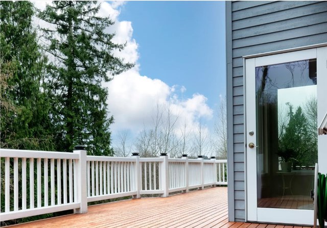 White deck with railing beside gray house and glass door under a blue sky