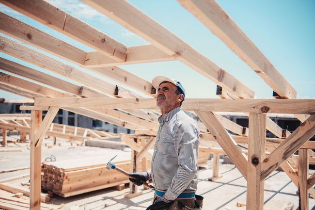 Construction worker under wooden roof framing at a building site, holding a tool and looking up