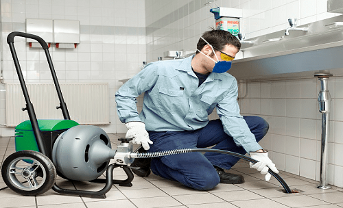 Funcionário com uniforme azul limpando um ralo de chão com uma mangueira em uma área de serviço revestida de azulejos
