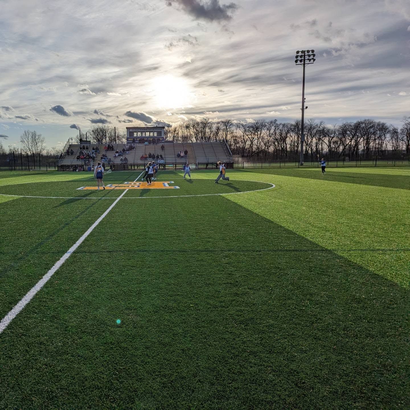 Soccer players on a green field at sunset, with bleachers and stadium lights in the background