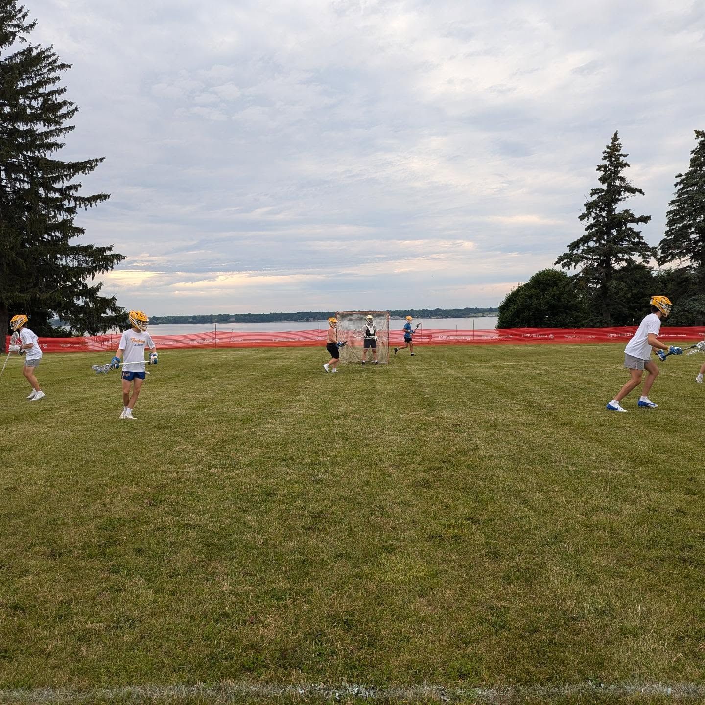Youth playing field game on grassy shore, with players in white and yellow near a red barrier under cloudy sky.