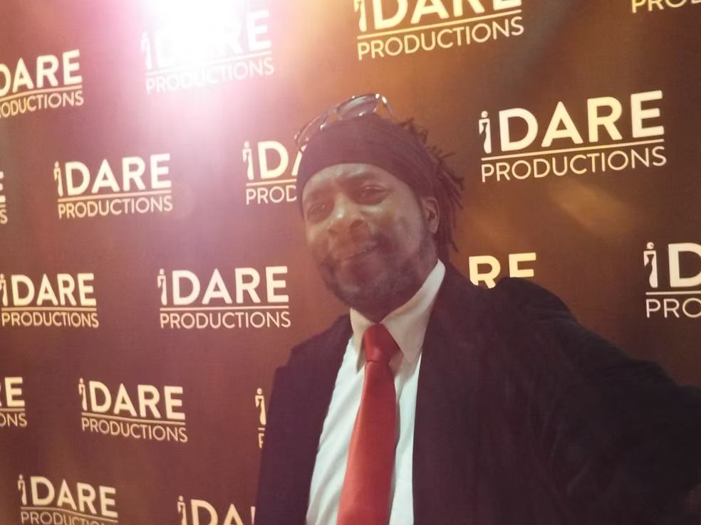 Man posing in a suit and red tie at a DARE Productions backdrop with warm lighting