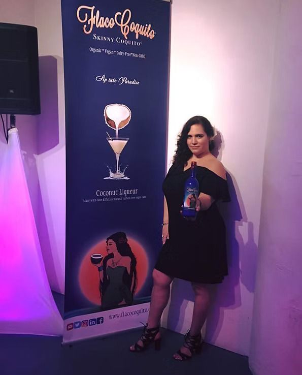 Woman in black dress posing beside a cocktail banner, holding a blue bottle under purple event lighting.