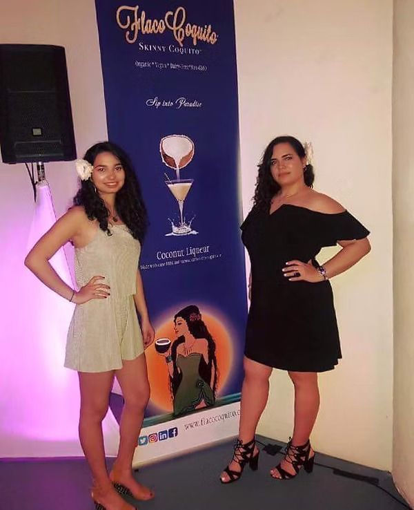 Two women posing beside a blue promotional banner in a hallway, wearing black and beige outfits.