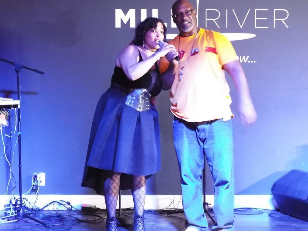 Two people posing on a stage with a microphone, in front of a purple 