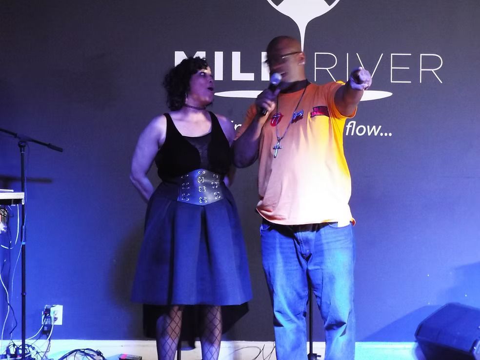Two performers onstage under blue and purple lights, one singing into a microphone beside a woman in a black dress.