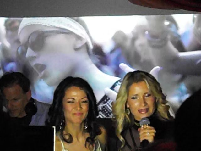 Two women singing into microphones on stage, with a projected black-and-white face in the background.