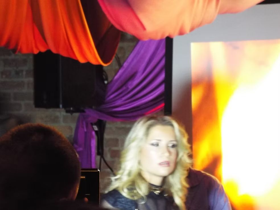 Blonde woman beside bright orange screen in a dim indoor venue with purple drapery