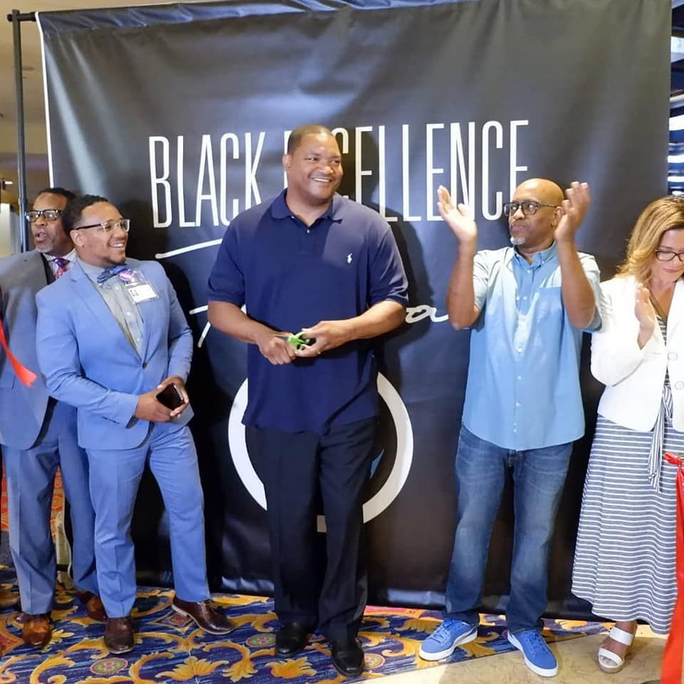 People standing in front of a BLACK EXCELLENCE banner, with one man speaking into a microphone. People standing in front of a BLACK EXCELLENCE banner, with one man speaking into a microphone.