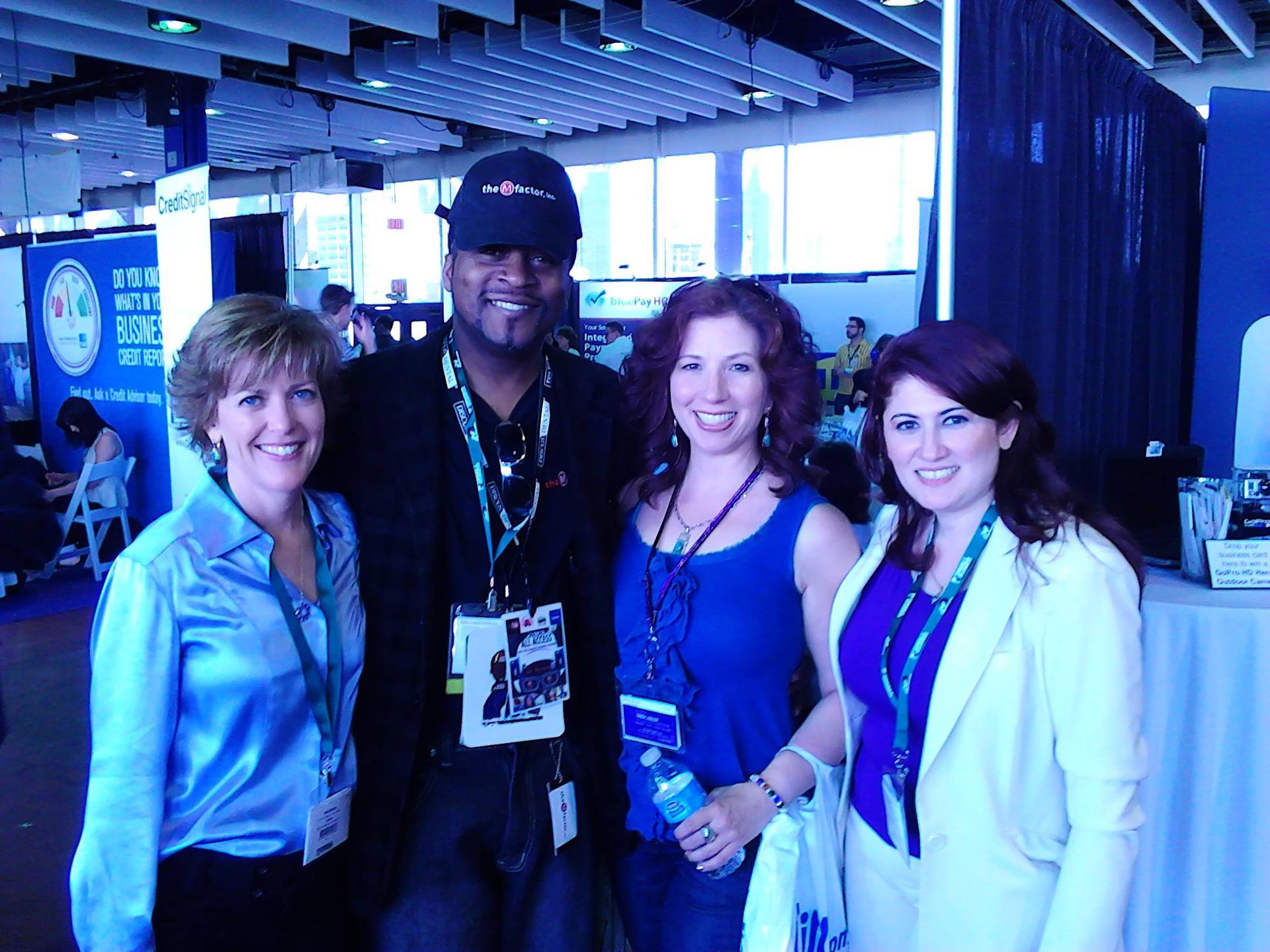 Four smiling people posing together in a blue-lit indoor venue, wearing lanyards and badges.