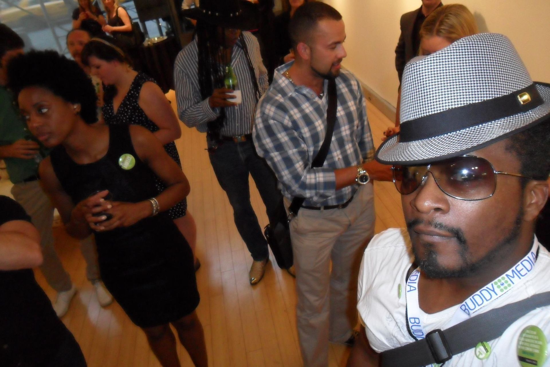 People mingling at an indoor event, with a man in a gray fedora and sunglasses in the foreground.