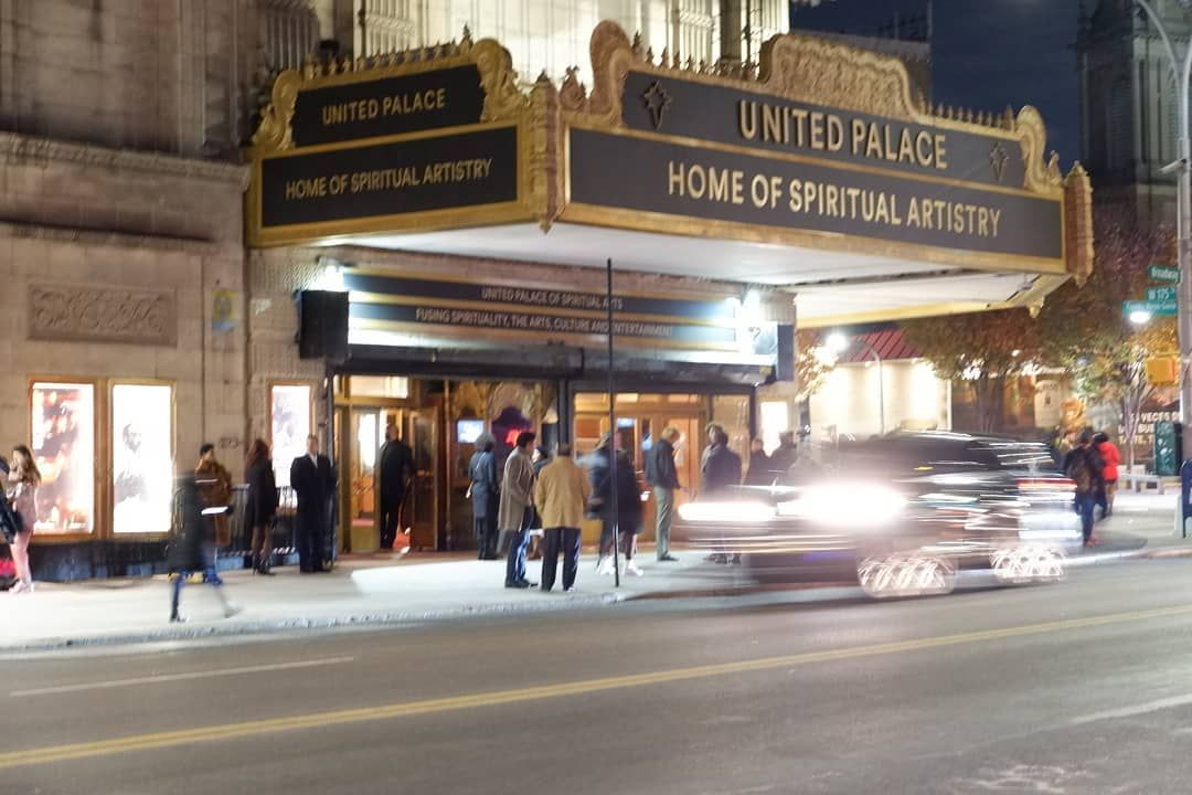 Nighttime entrance to United Palace theater with marquee lights, pedestrians, and a passing car on the street.