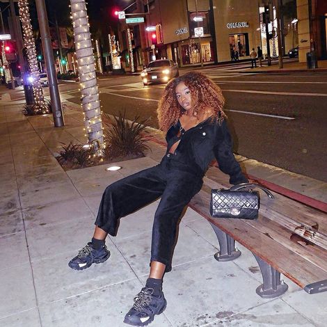 Person lounging on a bench at night on a city sidewalk, wearing black clothes and sneakers with a purse beside them.