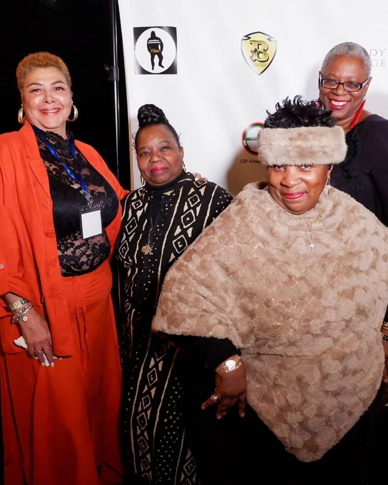 Four people smiling at a formal event, dressed in orange, black, and beige against a white backdrop.