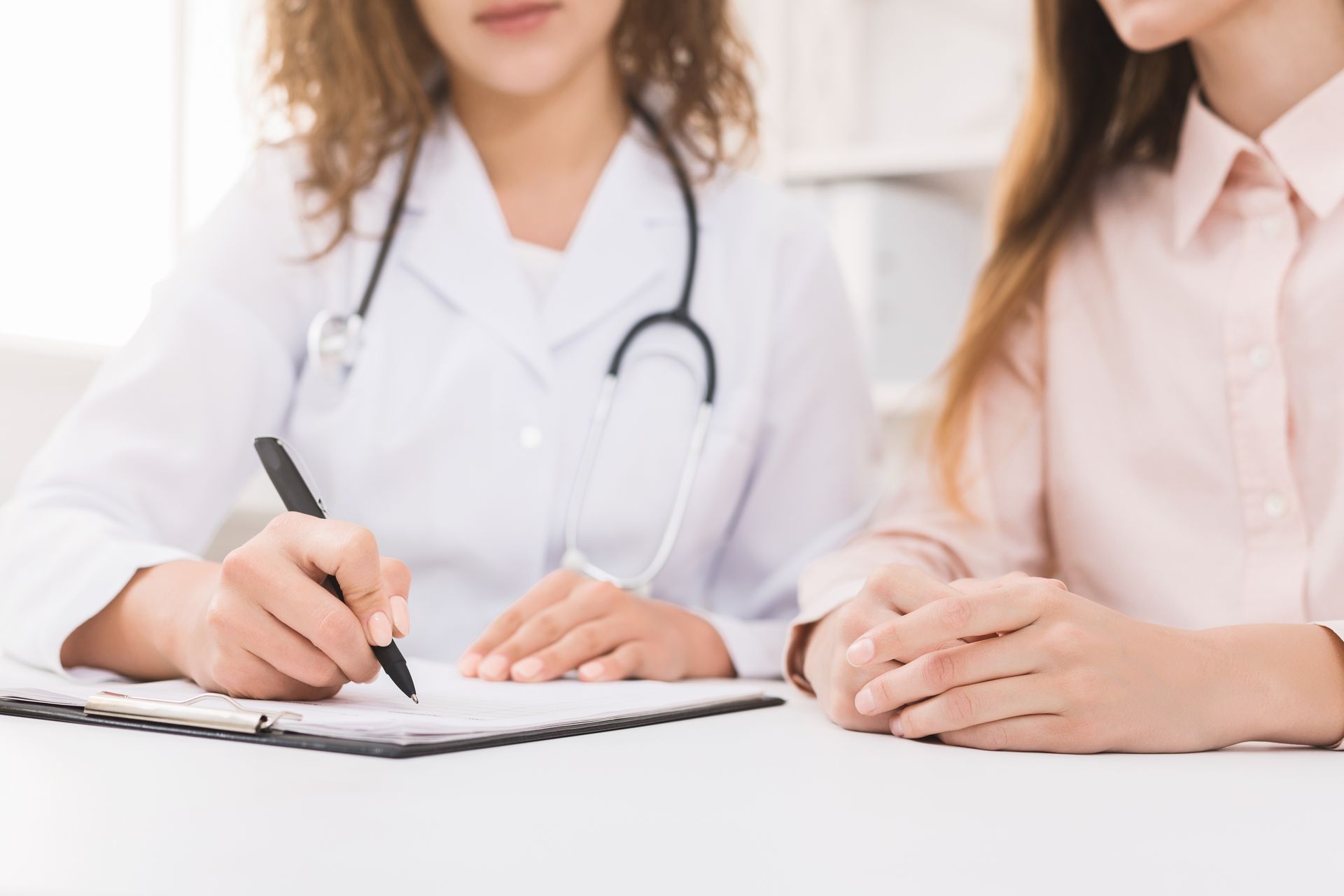 Healthcare professional wearing a white coat and stethoscope writing on a clipboard while sitting