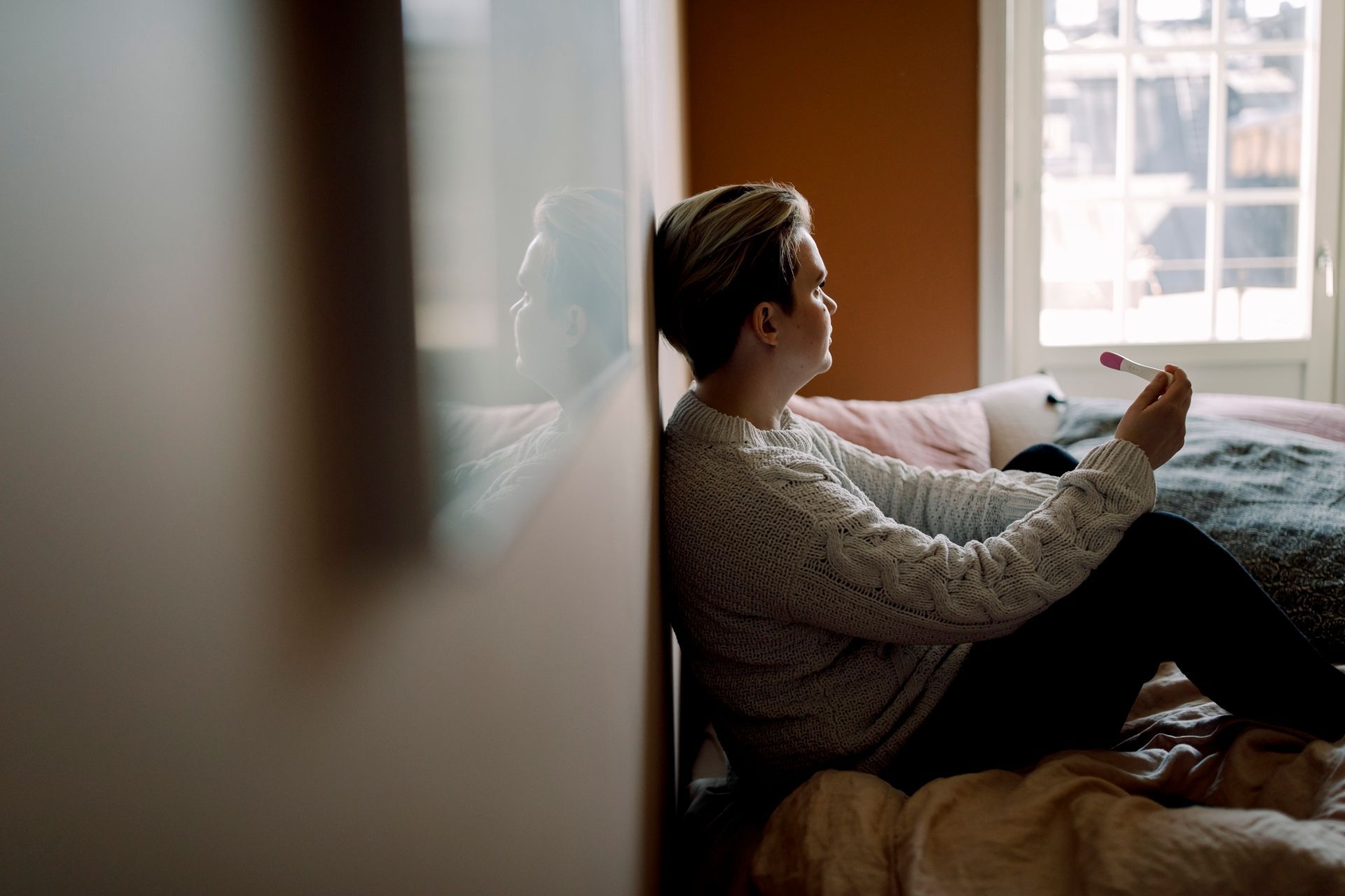Person sits on bed holding pregnancy test, leaning against wall.
