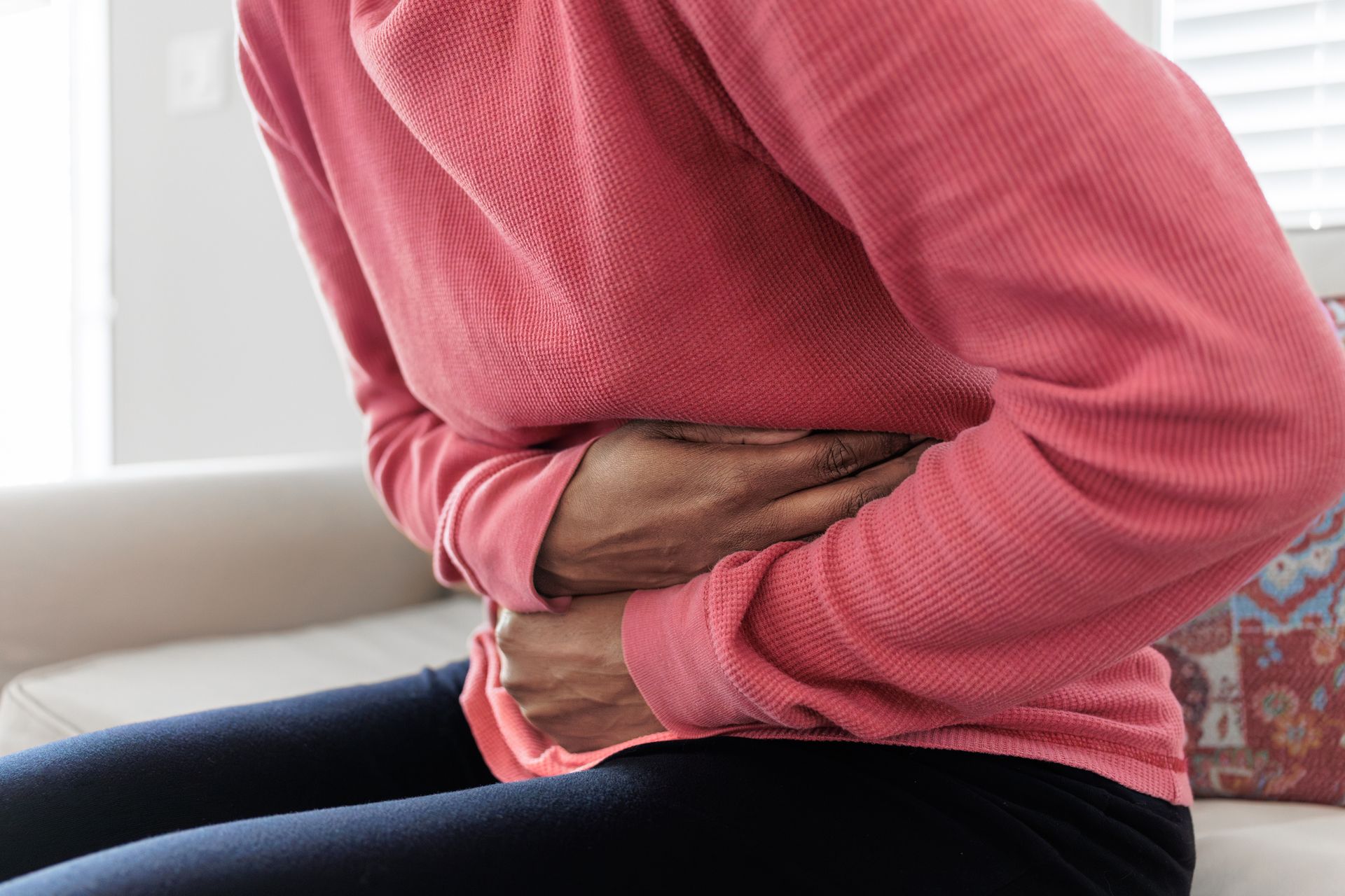Person sitting, holding stomach, possibly in pain; wearing pink shirt and dark pants.