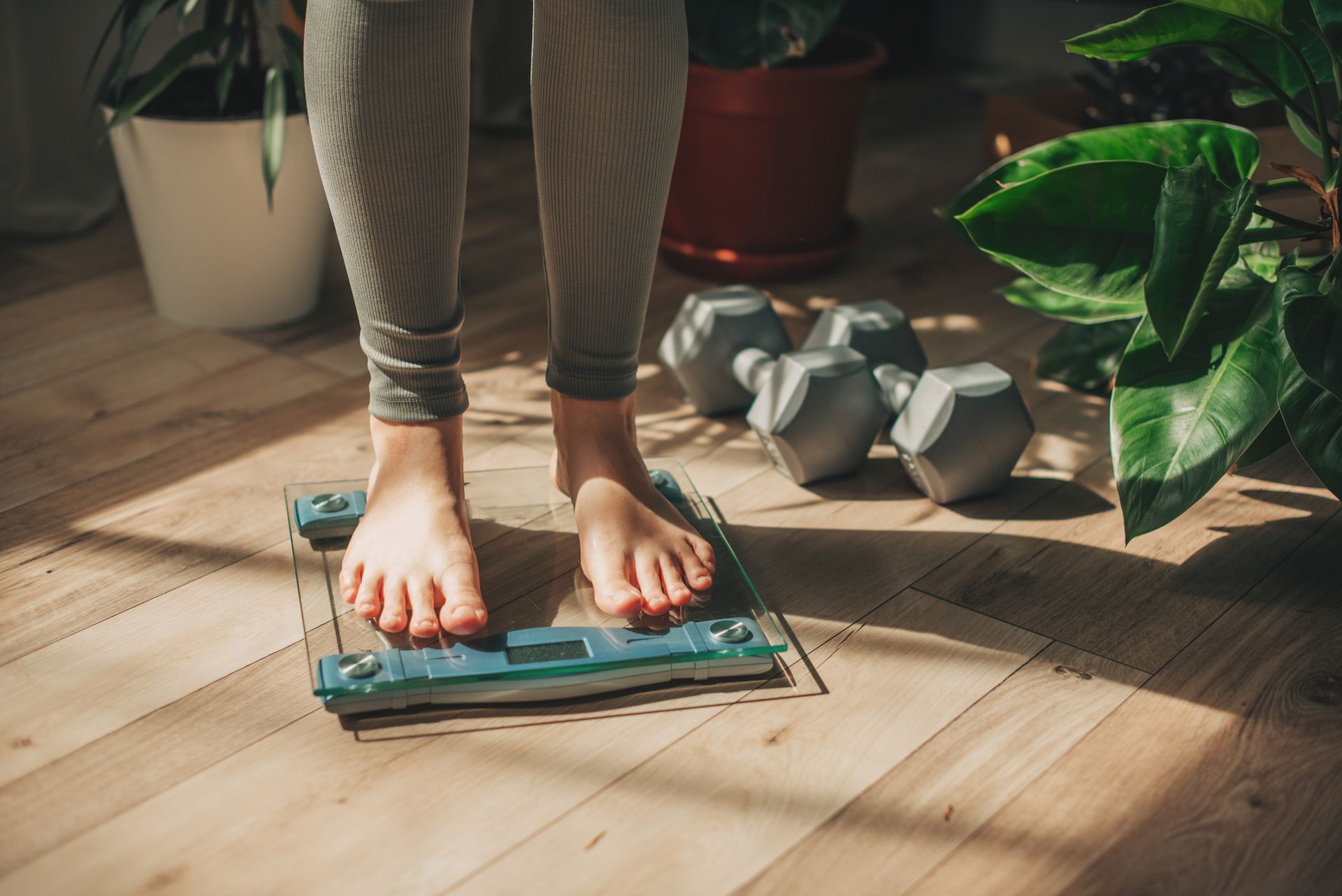 Person's feet on a scale, with dumbbells and plants in the background.