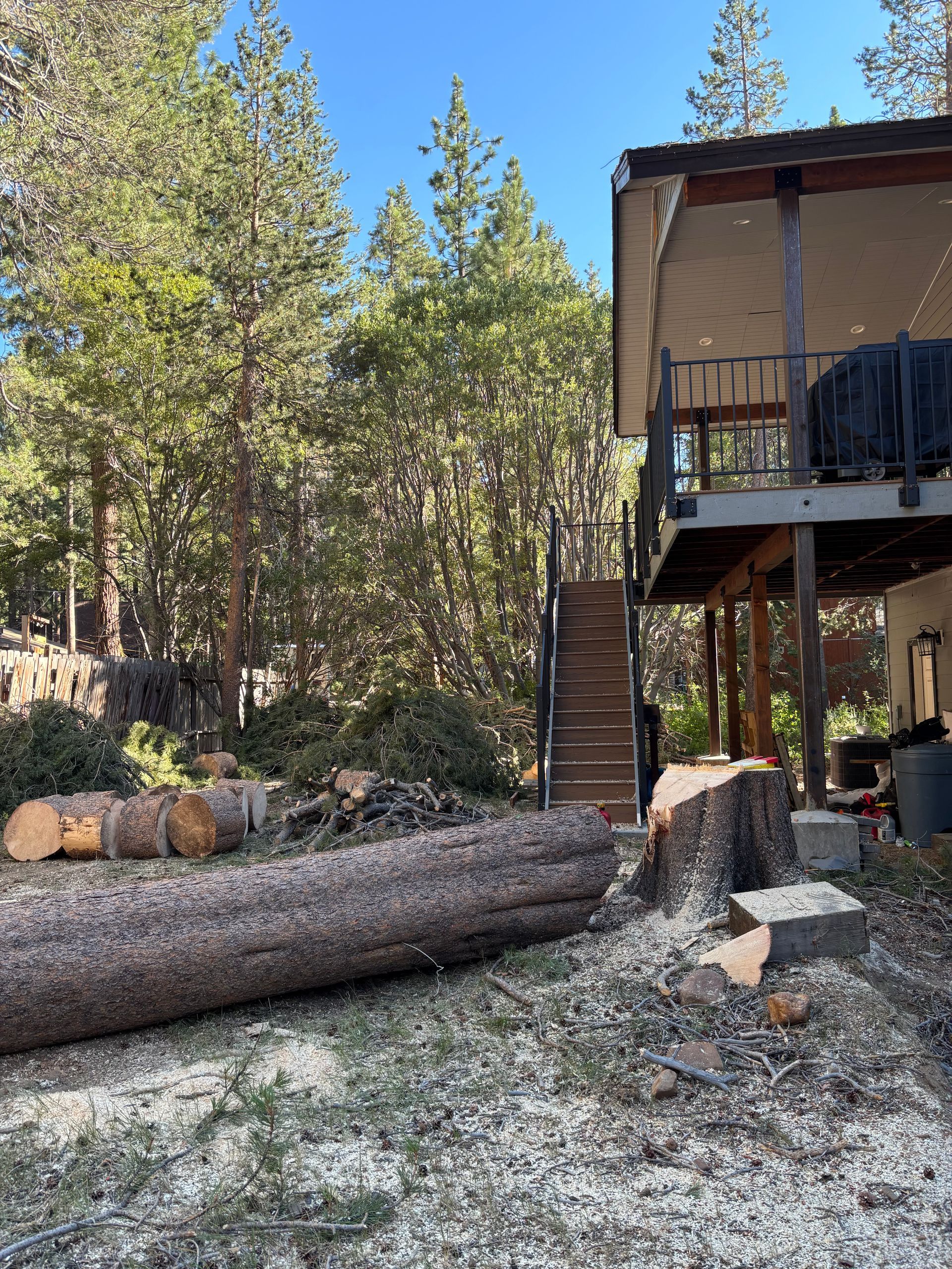 Logs and wood shavings near a home with a deck and stairs, set in a wooded area.