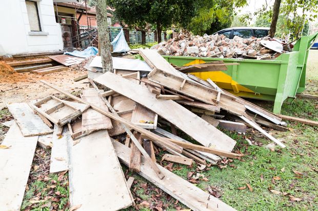 Pile of broken wooden boards and debris beside a green dumpster in a yard after demolition